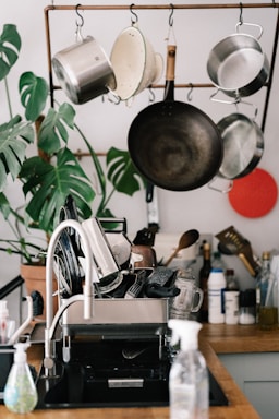 A kitchen setting features a dish rack filled with various clean kitchen utensils, including pots, pans, and glassware. Above the sink, a wooden rack holds hanging metal pots and pans. There are kitchen cleaning supplies on the counter, such as a dish soap dispenser. A green potted plant with large leaves adds a touch of nature to the scene.