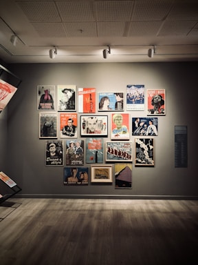 A gallery wall displaying a collection of vintage posters featuring various illustrations and textual elements. The posters are arranged in a grid-like pattern on a neutral-colored wall, each framed in either wood or a metallic finish. The floor is wooden, creating a warm, inviting ambiance.