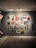 A gallery wall displaying a collection of vintage posters featuring various illustrations and textual elements. The posters are arranged in a grid-like pattern on a neutral-colored wall, each framed in either wood or a metallic finish. The floor is wooden, creating a warm, inviting ambiance.