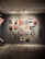 A gallery wall displaying a collection of vintage posters featuring various illustrations and textual elements. The posters are arranged in a grid-like pattern on a neutral-colored wall, each framed in either wood or a metallic finish. The floor is wooden, creating a warm, inviting ambiance.