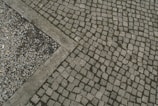 A paved area with cobblestones arranged in a geometric pattern, bordered by a small section of gravel. The arrangement and contrast between the cobblestones and gravel create an interesting visual texture.