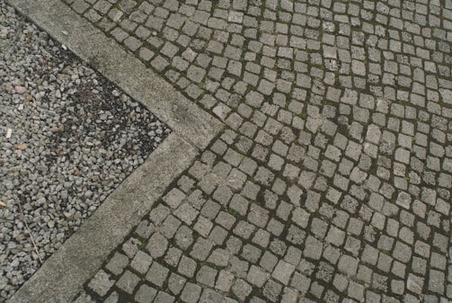 A paved area with cobblestones arranged in a geometric pattern, bordered by a small section of gravel. The arrangement and contrast between the cobblestones and gravel create an interesting visual texture.