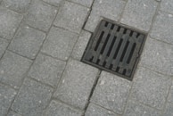 A grid-patterned sewer grate is set amid paving stones with a coarse texture. The grate is dark metal and contrasts with the gray paving stones surrounding it.