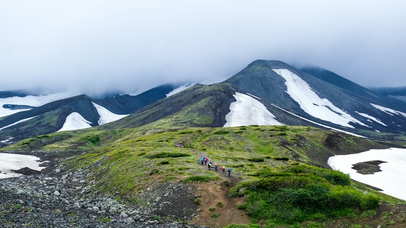 A mountain landscape features lush green hills with patches of snow and a group of hikers walking along a trail. The sky is overcast with low-hanging clouds that partially obscure the mountain peaks in the background.