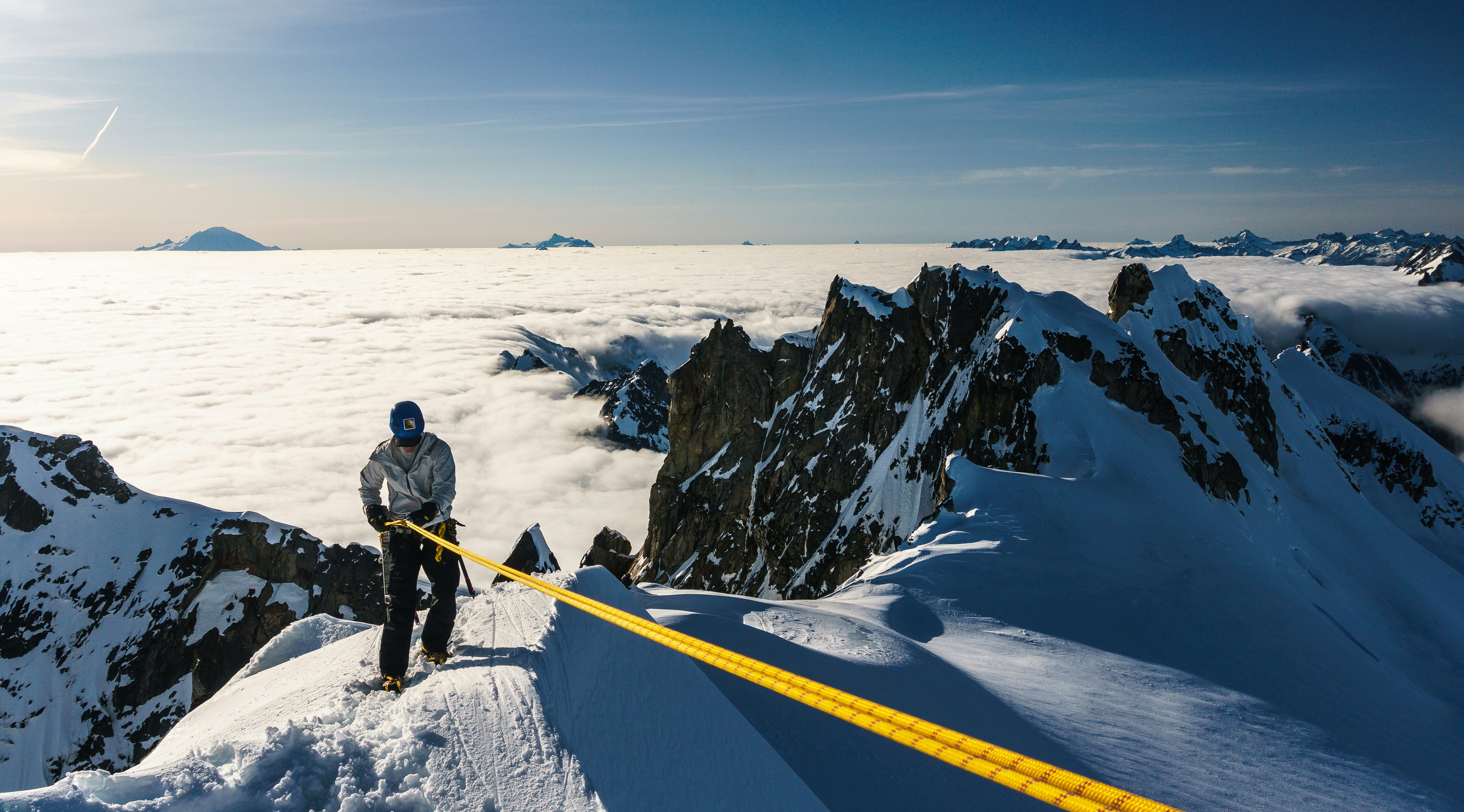 man in black jacket and black pants standing on snow covered mountain during daytime, Alpinist rappelling off a snowy mountain peak while above the clouds in the North Cascades National Park, Washington US