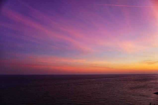 A serene ocean horizon with a vibrant sky displaying a gradient of purple, pink, and orange hues, reflecting the warm colors of a setting or rising sun.