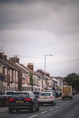 A residential street lined with brick houses, displaying cars parked on both sides and driving along the road. The sky is overcast, lending a muted tone to the scene. Each house is adorned with satellite dishes and chimneys, and a truck is visible further down the street.