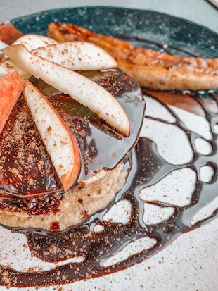 A dish featuring pancakes topped with sliced apples and drizzled with a rich, dark chocolate sauce. A dusting of brown spice, likely cinnamon, is sprinkled over the top. In the background, a caramelized banana adds a touch of contrast to the presentation.