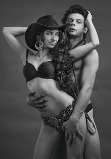 A black and white photograph featuring two individuals in an intimate pose. The woman is wearing a cowboy hat, a bra, and a patterned belt with a large circular ornament. The man is shirtless, holding the woman closely. Both appear confident and express strong body language.