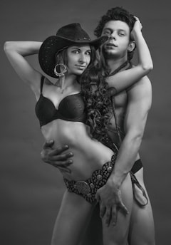 A black and white photograph featuring two individuals in an intimate pose. The woman is wearing a cowboy hat, a bra, and a patterned belt with a large circular ornament. The man is shirtless, holding the woman closely. Both appear confident and express strong body language.
