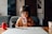 A young child is sitting at a table with a neutral expression, dressed in a brown jacket and drinking yogurt from a tetra pack using a straw. The table has a red toy robot and some other items placed on it. The background features a couch and wall art, creating a cozy, indoor setting.