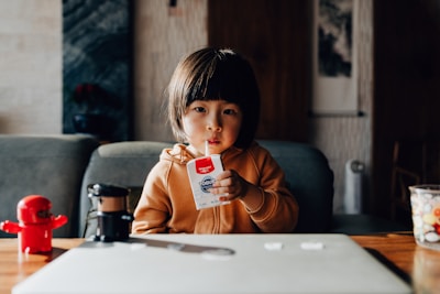 A young child is sitting at a table with a neutral expression, dressed in a brown jacket and drinking yogurt from a tetra pack using a straw. The table has a red toy robot and some other items placed on it. The background features a couch and wall art, creating a cozy, indoor setting.