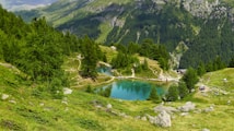 A picturesque alpine landscape featuring a serene, turquoise-colored lake surrounded by lush, green trees and rugged rocky terrain. Winding trails lead through the scene, with the backdrop of towering, forested mountains. A few people can be seen enjoying the natural beauty, engaging in activities such as hiking and resting.
