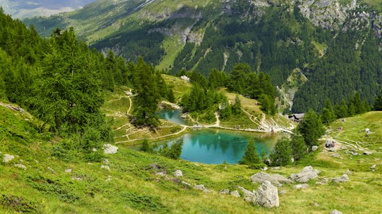 A picturesque alpine landscape featuring a serene, turquoise-colored lake surrounded by lush, green trees and rugged rocky terrain. Winding trails lead through the scene, with the backdrop of towering, forested mountains. A few people can be seen enjoying the natural beauty, engaging in activities such as hiking and resting.