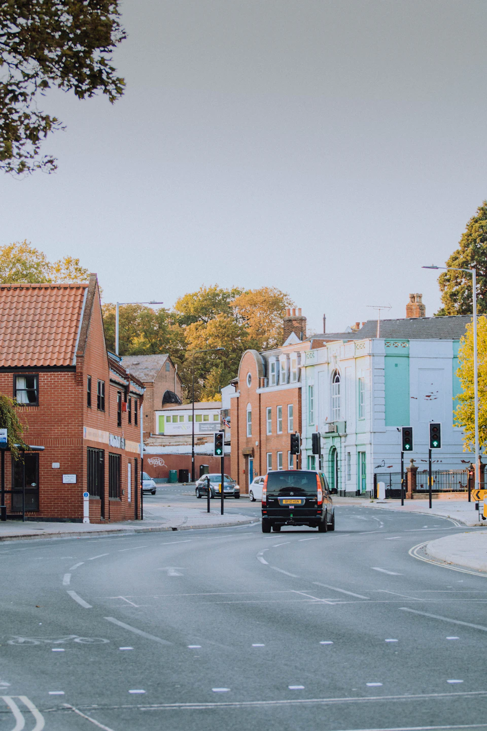 The image depicts a street scene with two-story brick buildings lining both sides, one on each side of the road. The buildings have red roofs and white walls, creating a harmonious color palette. A black car is driving down the street in front of these buildings, adding a sense of movement to the otherwise still scene. On either side of the street, there are trees with yellow leaves, suggesting that autumn has arrived.