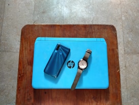 A blue laptop with an HP logo placed on a wooden surface. On top of the laptop, there is a blue smartphone and a wristwatch with a brown strap.