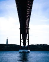 A view from beneath a large red suspension bridge spanning across a body of water, with a statue resembling Christ the Redeemer on the left perched atop a distant hill. The sky is mostly clear with a few clouds.