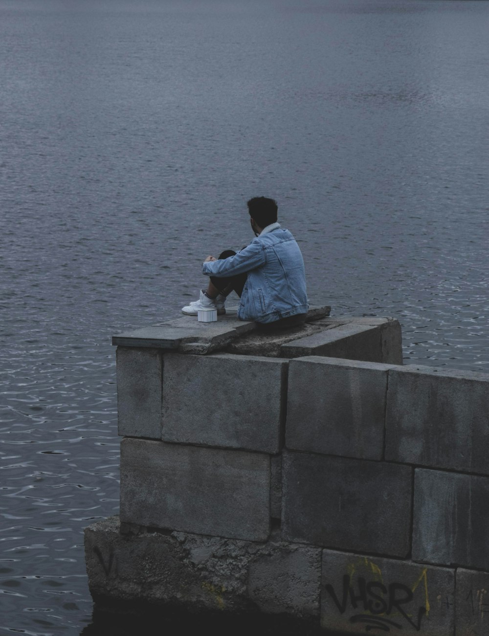 hombre con camisa de vestir blanca sentado en una pared de concreto cerca del cuerpo de agua durante el día
