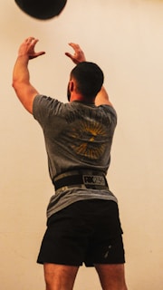 A man is exercising indoors with a medicine ball. He has both arms extended upward, appearing to throw the ball. He is wearing a gray t-shirt and black shorts, with a weightlifting belt around his waist.