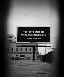 A black and white photograph features a billboard set on an urban street. The billboard bears the message, 'The truth can't die when journalism lives. inforum.news/truth'. Surrounding the billboard are urban structures, including a building and streetlights, with a moody, focused perspective leading to the board.