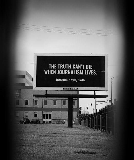 A black and white photograph features a billboard set on an urban street. The billboard bears the message, 'The truth can't die when journalism lives. inforum.news/truth'. Surrounding the billboard are urban structures, including a building and streetlights, with a moody, focused perspective leading to the board.