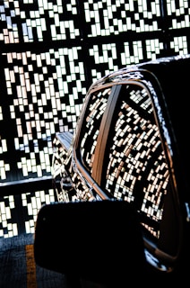 A shiny car is parked with an intricate pattern of small rectangular lights reflecting on its surface. The background features a decorative wall with geometric designs created by the same light pattern.