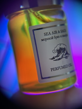 A close-up of a bottle labeled 'Sea Air & Sage' with a wave illustration. The contents appear to be a scented oil, and the bottle glows with colorful lighting, including shades of blue, purple, and orange.