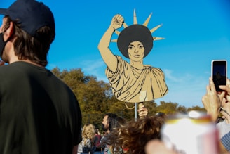 A crowd of people is gathered outdoors under a clear blue sky. A prominent cardboard cutout in the style of the Statue of Liberty is held aloft by an individual in the center, featuring an afro hairstyle. Several people in the foreground are visible, some taking photos with their phones.