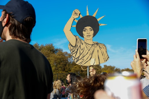 A crowd of people is gathered outdoors under a clear blue sky. A prominent cardboard cutout in the style of the Statue of Liberty is held aloft by an individual in the center, featuring an afro hairstyle. Several people in the foreground are visible, some taking photos with their phones.