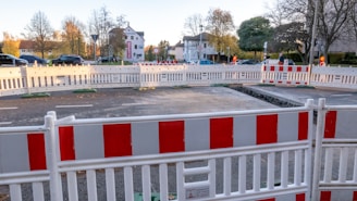A construction site is cordoned off with red and white barricades on a road. Cars are parked in the background, and there are trees with autumn foliage. A partly dug trench is visible inside the barricaded area. Buildings and walking pedestrians can be seen behind the enclosure.