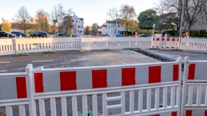 A construction site is cordoned off with red and white barricades on a road. Cars are parked in the background, and there are trees with autumn foliage. A partly dug trench is visible inside the barricaded area. Buildings and walking pedestrians can be seen behind the enclosure.