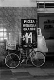 A black and white image features a bicycle parked in front of a tiled wall. There is a sign advertising 'Pizza Muzzarella Grande' for $14, with delivery options, placed beside the bicycle. The shop interior is partially visible with various kitchen items on shelves.