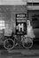 A black and white image features a bicycle parked in front of a tiled wall. There is a sign advertising 'Pizza Muzzarella Grande' for $14, with delivery options, placed beside the bicycle. The shop interior is partially visible with various kitchen items on shelves.