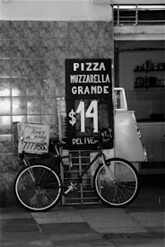 A black and white image features a bicycle parked in front of a tiled wall. There is a sign advertising 'Pizza Muzzarella Grande' for $14, with delivery options, placed beside the bicycle. The shop interior is partially visible with various kitchen items on shelves.