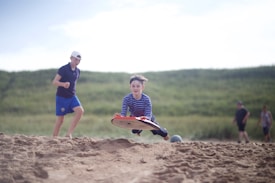 A young person rides a sandboard across a sandy surface, displaying an energetic action shot. Another person in the background is running and wearing athletic shorts and a hat, while additional figures are visible in the distance. Lush green grass fills the background, providing a natural setting.