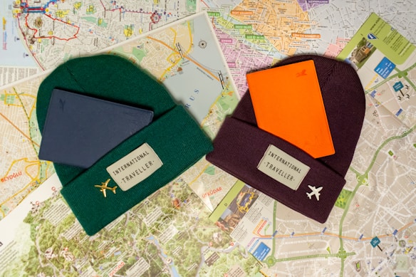 Two beanies labeled 'International Traveller' are placed on top of various city maps. One beanie is dark green with a gold airplane pin and has a blue notebook on it. The other is maroon with a white airplane pin and has an orange notebook on it. The background consists of detailed street maps with different colors and markings.