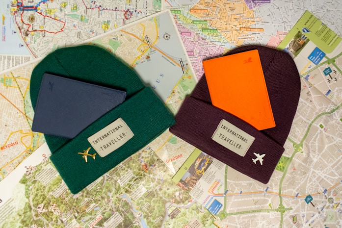 Two beanies labeled 'International Traveller' are placed on top of various city maps. One beanie is dark green with a gold airplane pin and has a blue notebook on it. The other is maroon with a white airplane pin and has an orange notebook on it. The background consists of detailed street maps with different colors and markings.