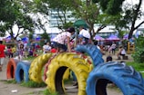 Children and adults are enjoying a lively playground. Kids are climbing on colorful tires arranged as play equipment. The scene is set in a park with trees, greenery, and various play structures in the background.