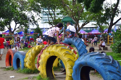 Children and adults are enjoying a lively playground. Kids are climbing on colorful tires arranged as play equipment. The scene is set in a park with trees, greenery, and various play structures in the background.