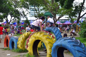 Children and adults are enjoying a lively playground. Kids are climbing on colorful tires arranged as play equipment. The scene is set in a park with trees, greenery, and various play structures in the background.
