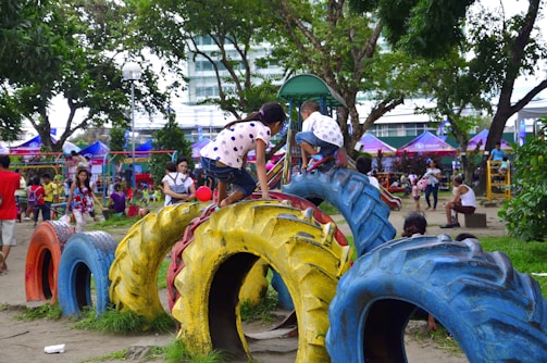 Children and adults are enjoying a lively playground. Kids are climbing on colorful tires arranged as play equipment. The scene is set in a park with trees, greenery, and various play structures in the background.