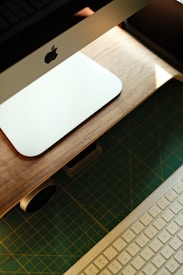 An Apple computer with a wooden stand is positioned on a cutting mat. A wireless keyboard is visible in the foreground, and a small horizontal device is placed beneath the computer.