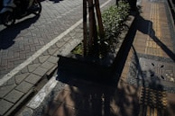 A paved pedestrian walkway with tactile paving for visually impaired individuals runs alongside a road where a scooter is visible. There is a small section of greenery with plants in a raised concrete bed supported by wooden stakes. The walkway features a decorative metal grate and is shadowed by nearby objects, creating an interplay of light and dark patterns.