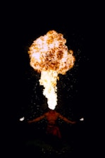 A person is performing a fire-breathing act, expelling a large plume of fire from their mouth while holding lit torches in both hands. The background is dark, making the bright flames stand out vividly.