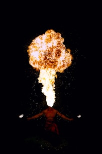 A person is performing a fire-breathing act, expelling a large plume of fire from their mouth while holding lit torches in both hands. The background is dark, making the bright flames stand out vividly.