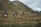 A series of small red cabins with triangular roofs are set against a backdrop of rolling green hills and mountains. The landscape is lush with patches of vegetation and a cloudy sky overhead.