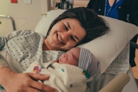 A woman lies in a hospital bed smiling joyfully while holding a newborn baby wrapped in a blanket. The setting appears to be a hospital room, indicated by her hospital gown and the IV line visible on her hand.