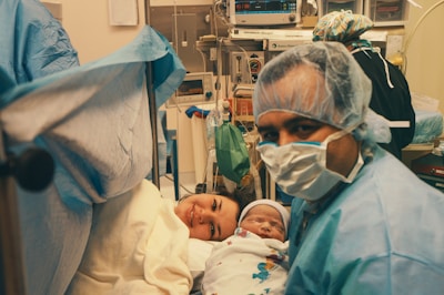A medical setting with a new mother lying on a hospital bed, smiling while holding her newborn baby. A person, likely a medical professional, is wearing a surgical mask and gown, positioned near the mother. Medical equipment is visible in the background, indicating a hospital room or operating theater.