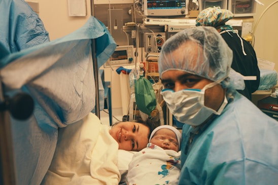 A medical setting with a new mother lying on a hospital bed, smiling while holding her newborn baby. A person, likely a medical professional, is wearing a surgical mask and gown, positioned near the mother. Medical equipment is visible in the background, indicating a hospital room or operating theater.