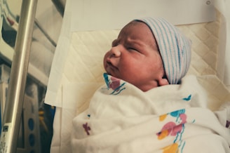 A newborn baby is wrapped in a white hospital blanket with colorful patterns. The baby is wearing a striped beanie and is lying on a padded surface.